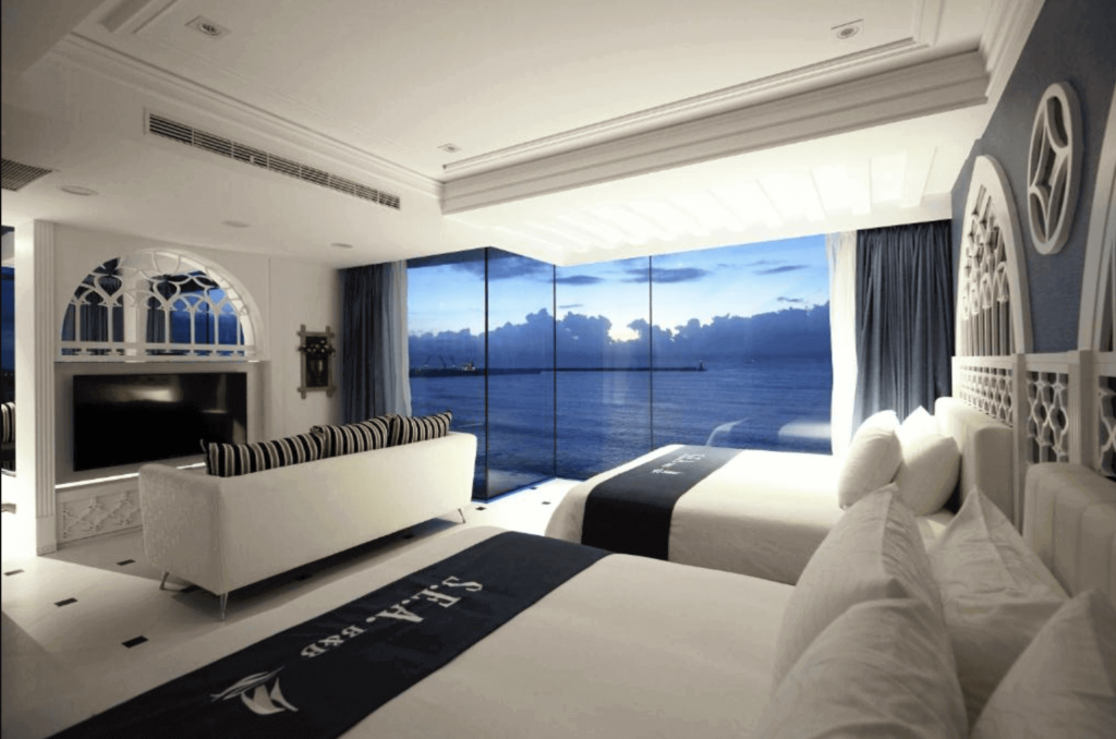 depicts a luxurious hotel room with a nautical theme, evident in the white and navy blue color scheme. The room features two twin beds with stylized nautical decor and a comfortable sitting area with a black-and-white striped sofa facing a flat-screen TV. An ornate, white Gothic-style window complements the room's elegant design. The room boasts a panoramic view of the ocean and skyline through expansive floor-to-ceiling windows, allowing natural light to flood the space and highlight the intricate design details.