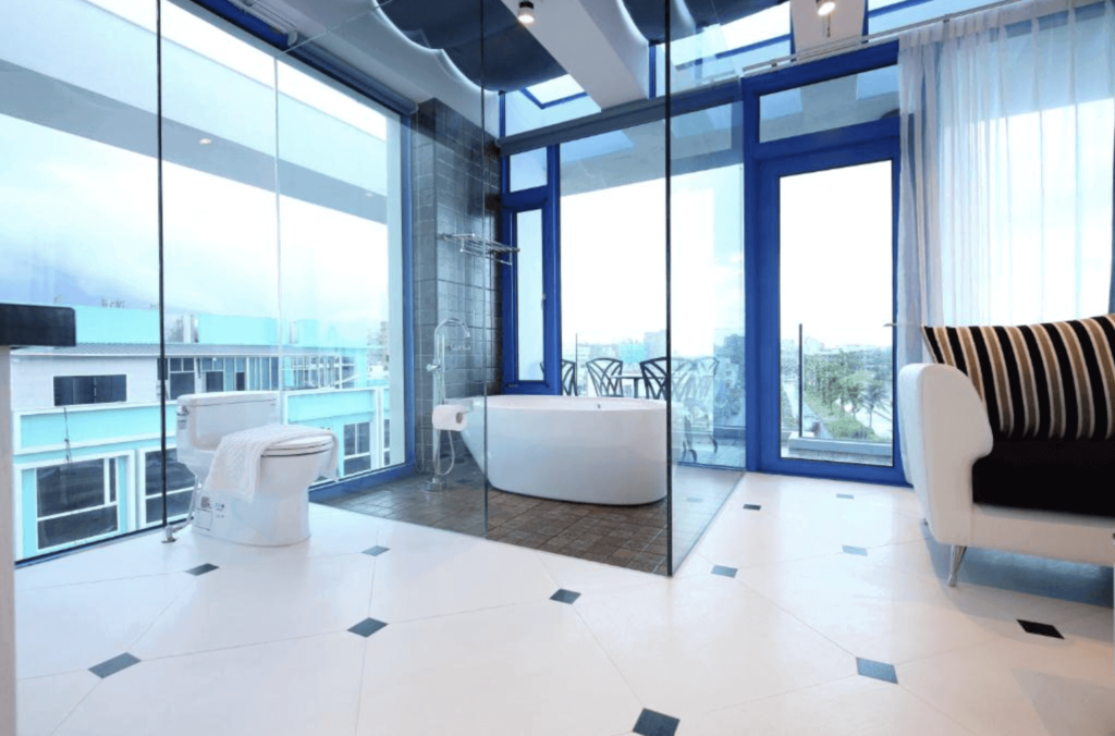 showcases a bright and modern hotel bathroom with striking blue accents. It has a freestanding bathtub and a separate glass-enclosed shower area, both positioned to offer views of the outdoors through large, floor-to-ceiling windows. The interior is sleek with white walls and tiles accented with blue details, and a black-and-white striped sofa adds a chic touch. The outdoor view is partially obscured but suggests a high-rise vantage point over an urban area.