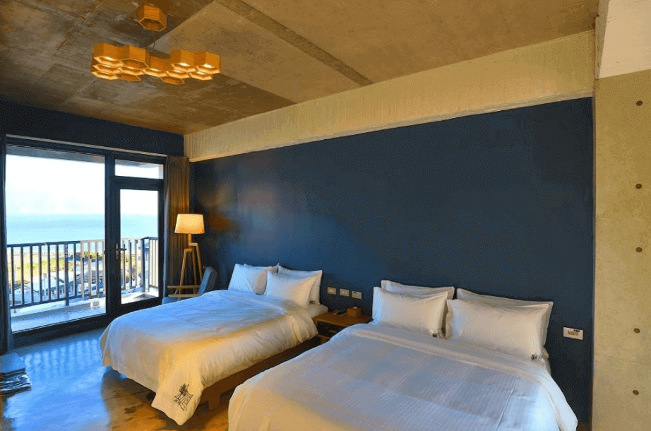 Modern beachfront hotel room with two beds, concrete ceiling, and a large window offering an expansive sea view