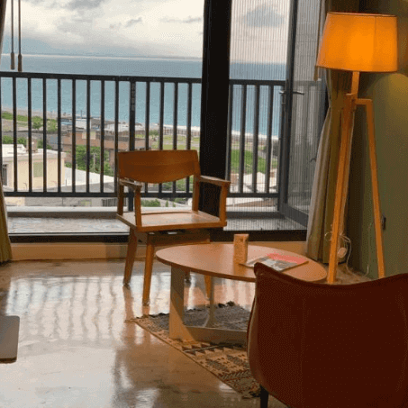 Hotel room corner with ocean view, featuring a stylish armchair, wooden table, and a floor lamp, creating a cozy reading nook.