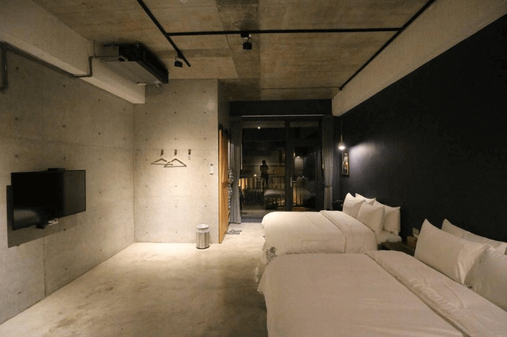 【花蓮市海景住宿】七星潭泥作輕旅民宿的夜間房間以簡約的工業風設計為主，牆面裸露水泥質感，搭配昏黃的燈光營造放鬆的入住氛圍，雙床設置適合多人入住，窗外可通往陽台欣賞夜景。
