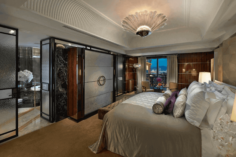 【台北親子飯店】台北文華東方的酒店套房的圖片，房間內部裝潢典雅，具有現代化設計元素。大床上鋪有多層枕頭和柔軟的床單，床頭有壁燈照明。房間的牆壁和門框使用了深色木材與玻璃結合的設計，增添了質感。天花板上裝有一個華麗的吊燈，整體氛圍非常舒適且高雅。