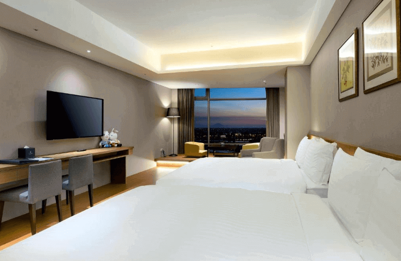Spacious hotel room with ambient lighting, cityscape view through large windows, twin beds with white linens, desk area, flat-screen TV, and decorative plush toys