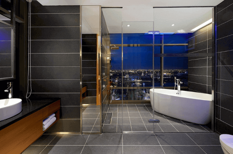 Hotel bathroom with floor-to-ceiling windows offering a night view of the city, featuring a walk-in shower, modern freestanding bathtub, sleek black tiles, and well-lit vanity area