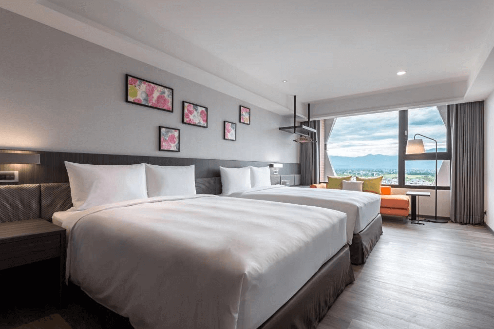 【宜蘭溫泉飯店】宜蘭館煙波大飯店的雙床客房，兩張大床整齊擺放，牆上裝飾有花卉圖案的掛畫。房間右側有一個大窗戶，窗外可以看到遼闊的山景和天際線，窗邊設有一張橘色的沙發和抱枕，營造出舒適的氛圍。