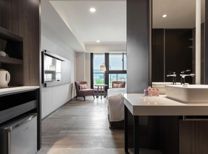 Sleek hotel room kitchenette with modern appliances, flat-screen TV, minimalist decor, and an open-plan layout leading to a cozy bedroom with mountain views