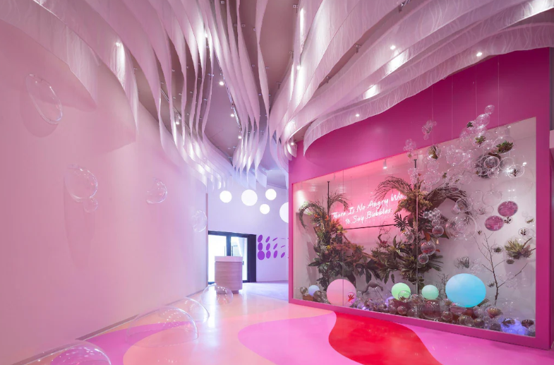 Futuristic hotel interior with whimsical bubble-inspired design, vibrant pink and purple tones, dynamic lighting, and a decorative botanical installation with a motivational quote