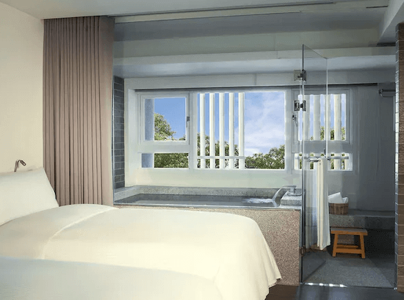 【宜蘭溫泉飯店】捷絲旅宜蘭礁溪館的一間配有大床的房間，床旁設有一個浴缸，位於落地窗旁邊，讓住客在泡澡時可欣賞窗外的美麗景色。房間的設計簡潔，白色牆面搭配大窗，整體氛圍舒適且充滿自然光。