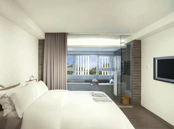 Contemporary hotel bedroom with a large bed, crisp white linens, flat-screen TV, and a unique transparent glass bathroom wall with privacy curtains