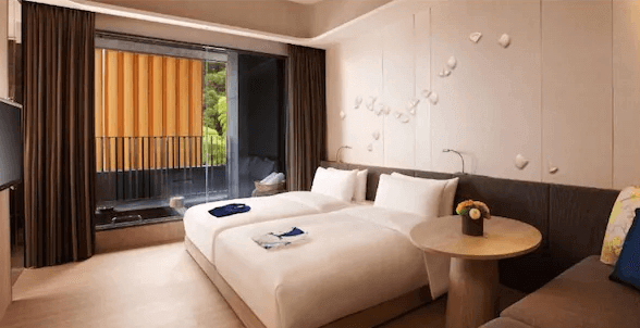 【宜蘭溫泉飯店】晶泉丰旅的一間現代溫馨的雙床房，房間內的床鋪舒適，旁邊有一個小圓桌和花卉裝飾。房間設有落地窗，通往一個陽台，陽台內有私人浴池，讓住客可以在戶外享受泡湯的樂趣，背後的山景令人放鬆。