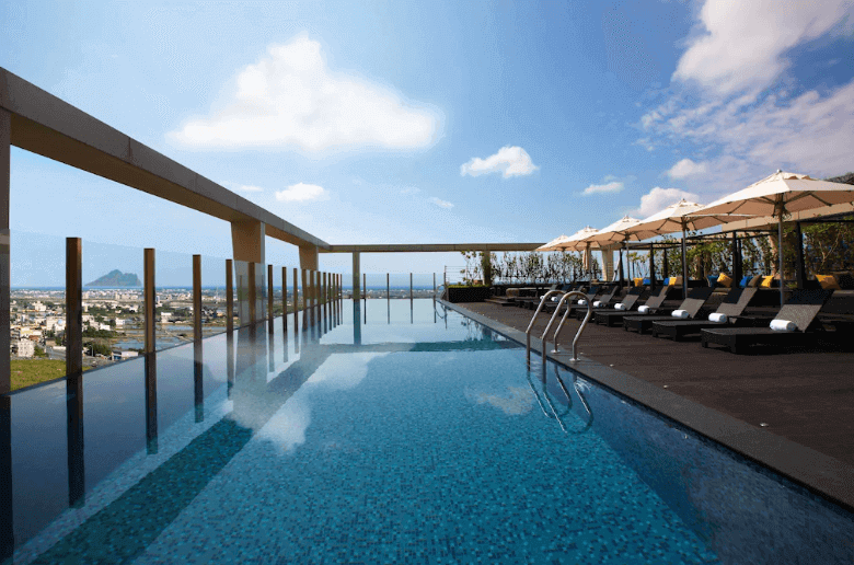Luxurious hotel rooftop infinity pool with panoramic views of the city skyline, sun loungers under umbrellas, clear blue skies, and a serene atmosphere for relaxation