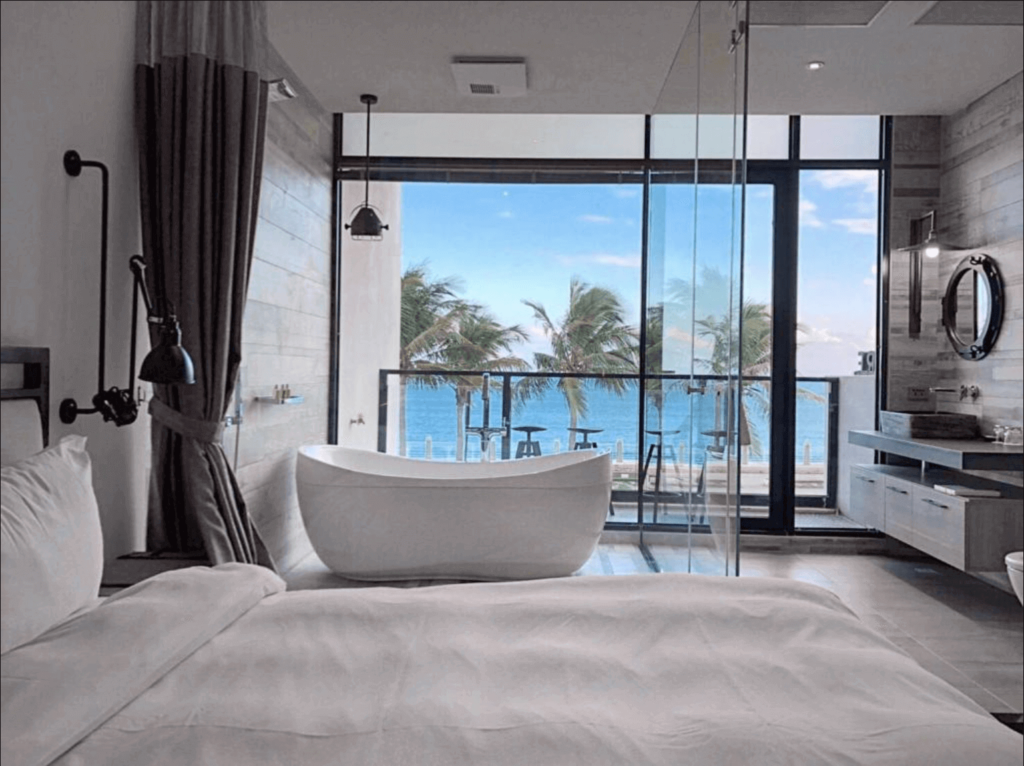 uxurious hotel room with a large, comfortable bed in the foreground. The room features a modern, minimalist aesthetic with neutral colors. A glass partition separates the sleeping area from the ensuite bathroom, which includes a standalone bathtub. Through the large glass windows, one can admire a beautiful ocean view with palm trees and a clear blue sky.