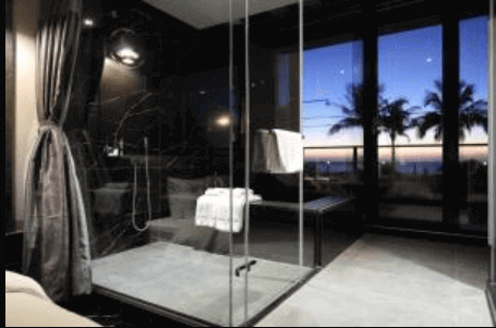 taken from inside a hotel room during the evening. The room overlooks a serene beach, visible through floor-to-ceiling windows. The room is elegantly designed with a dark color scheme that contrasts with the dusk light outside. The interior includes a reflective glass shower stall that mirrors the tranquil beach scene, enhancing the room's peaceful ambiance.