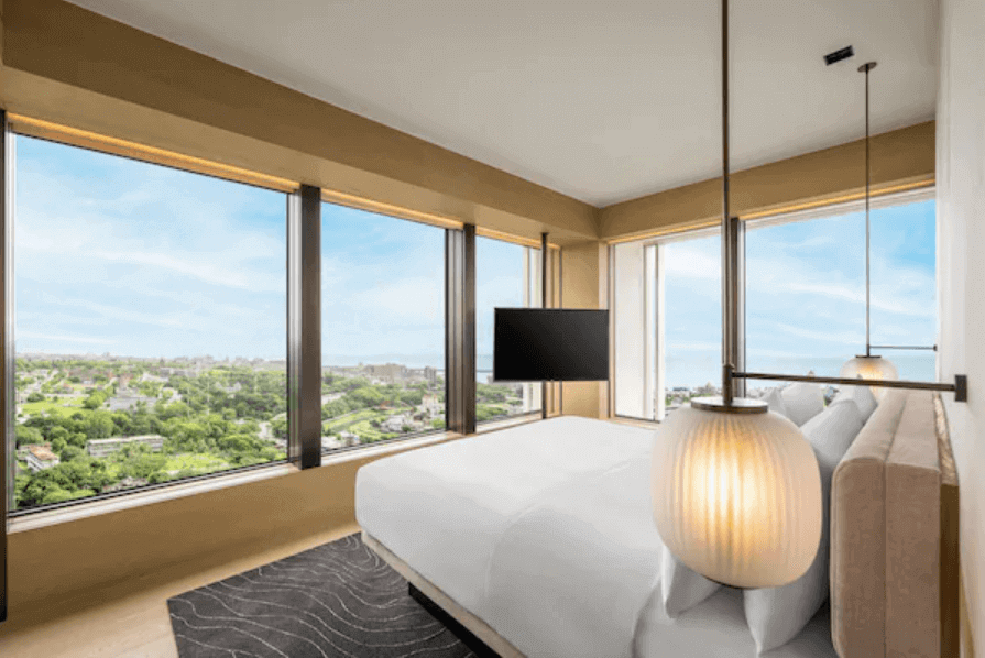 features a sophisticated hotel room with a panoramic city view through large, corner windows. The room is decorated with warm beige tones, and the bed is dressed in pristine white bedding. To the side, a stylish pendant light hangs above a nightstand, while a modern flat-screen TV is mounted opposite the bed. The design is elegant yet inviting, emphasizing comfort and the scenic urban landscape outside.