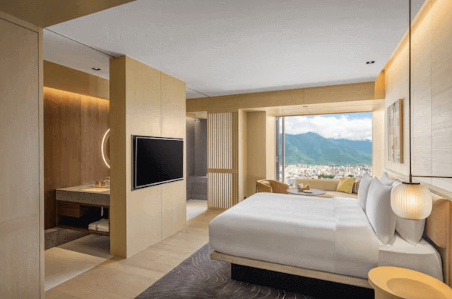 is another angle of the hotel room, highlighting a king-sized bed facing a window that boasts a breathtaking view of a mountainous landscape. A flat-screen TV is positioned within a partition wall, which also serves as a vanity with a round mirror on the opposite side. The room’s decor features a harmonious blend of neutral tones and natural light, creating a relaxing and luxurious environment.