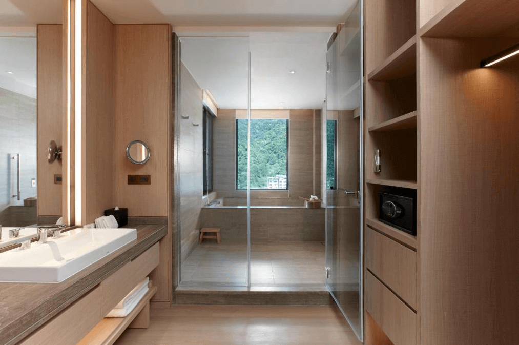 【宜蘭溫泉飯店】礁溪寒沐酒店的現代化的浴室，內部以淡木色為主，設有寬敞的洗手台和鏡子，旁邊是一個寬敞的淋浴間和浴缸區。浴缸區的大窗戶提供了自然光，窗外可見綠意盎然的山景。