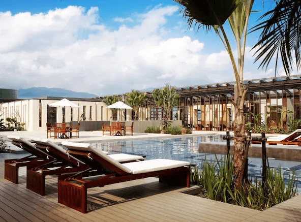 【宜蘭溫泉飯店】蘭城晶英酒店的戶外游泳池區，背景是晴朗的天空和遠山景色。池邊擺放著幾張躺椅供住客放鬆。整個戶外區域設計時尚，配有木質甲板和綠植，營造出度假村的氛圍。