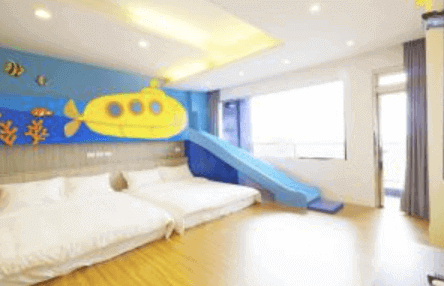 This picture shows a playful and colorful children's bedroom within a hotel. The room features a large bed and a smaller bed with bright blue sheets, both set against a mural of an underwater scene with fish and a submarine. A blue slide leads down from the main bed, adding a fun element for kids. The room is designed to spark imagination and play, with ample space for children to move around and enjoy.