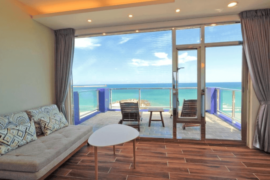 【花蓮市海景住宿】貳館海景民宿的客廳，設有寬大的玻璃窗戶和門，讓住客可以享受無敵的海景。客廳內部裝潢簡約，擺放著舒適的沙發和小茶几，地板採用木質材料，營造出溫馨的氛圍。窗外的陽台上設有休閒椅，讓住客能夠在戶外享受海風和美景，適合放鬆和休憩