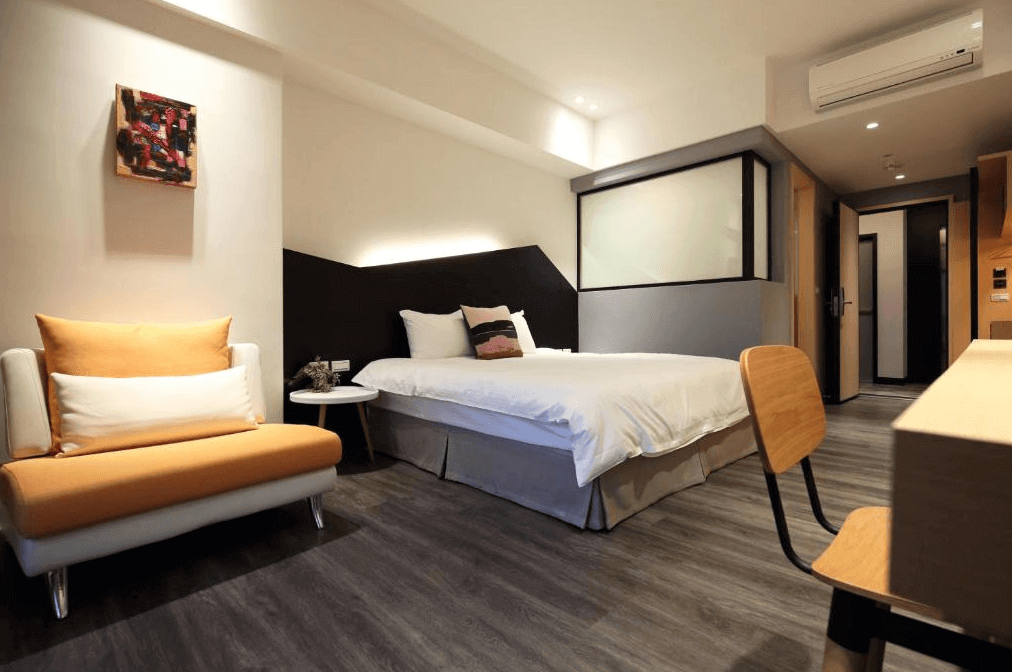 【花蓮市海景住宿】那山吶海‧海森威人文旅宿的臥室，設有一張大床，床上鋪著白色床單並配有舒適的枕頭。房間內的牆面裝飾著一幅抽象畫，增添了藝術氣息。床邊有一張橙色和白色相間的沙發，旁邊擺放著一個小圓桌，營造出舒適的休憩角落。房間內部採用木質地板，增添了溫暖的氛圍。書桌和椅子設置在房間的一角，提供了工作和學習的空間。整體設計簡約而時尚，適合追求舒適與現代感的住客。