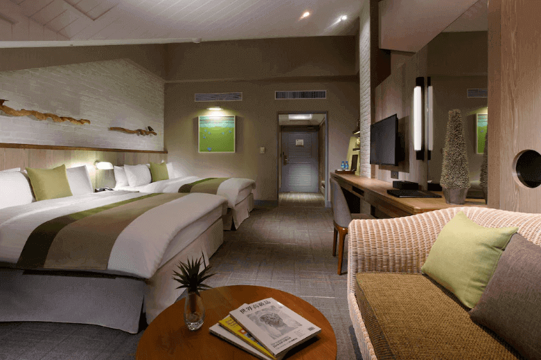 "台北親子飯店"【陽明山親子住宿】陽明山天籟渡假酒店的客房，配有兩張雙人床，床鋪上鋪有白色床單和綠色裝飾布，並搭配多個靠枕。房間內設有木質書桌、平面電視、舒適的沙發座椅和小圓桌，上面擺放著雜誌和小型盆栽。牆上裝飾著藝術畫作和木質擺設，燈光柔和，營造出溫暖舒適的氛圍。