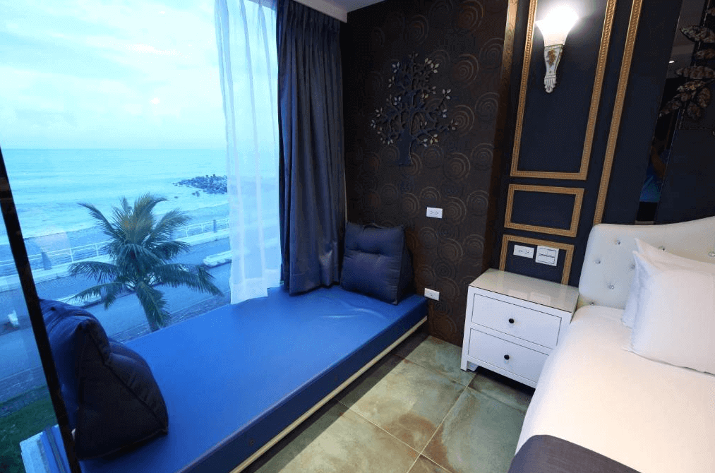 【花蓮市海景住宿】舒適的臥室一角，設有一個靠窗的休息區，可以俯瞰到棕櫚樹和海景。房間採用藍色和金色的裝飾風格，床旁設有白色的床頭櫃，營造出高雅的住宿環境