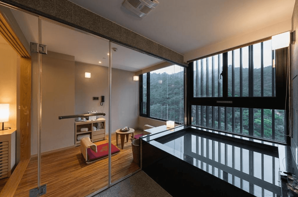 北投亞太飯店溫泉浴室：一間設有大型落地窗的溫泉浴室，窗外可見綠意盎然的山景。浴室內設有一個黑色石製浴缸，旁邊有休閒區，提供毛巾和其他浴室用品，營造出放鬆的氛圍