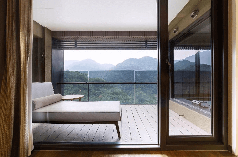 "台灣最貴飯店"北投麗禧溫泉酒店陽台景觀：從房間內看向陽台，陽台設有舒適的沙發和圓形茶几，背景是壯麗的山景和森林。玻璃門讓室內和室外的景色融為一體，提供寧靜的休閒空間。