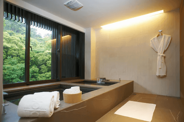 "台灣最貴飯店"北投麗禧溫泉酒店私人浴室：一間設有大窗戶的現代化私人浴室，窗外可見茂密的綠色植被。浴室內有一個大型浴缸，旁邊放置了浴巾和浴袍，營造出放鬆和舒適的氛圍