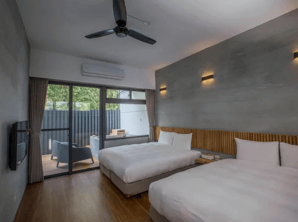 山下的厝溫泉民宿的雙人床房，房內有兩張雙人床，牆壁採用灰色水泥質感，整體設計現代簡約。房間內設有大窗戶和陽台，可以通往戶外，提供充足的自然光和休息空間。