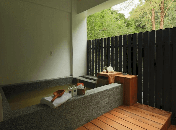 山下的厝溫泉民宿的私人溫泉區，設有石製浴缸和木製地板。周圍圍有木柵欄，提供隱私保護。浴缸旁放有毛巾和洗浴用品，背景是綠意盎然的戶外景色，讓住客能夠享受放鬆的泡澡時光。