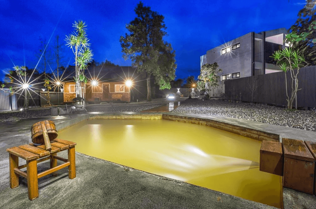山下的厝溫泉民宿的戶外溫泉池，夜晚拍攝，池水呈現天然的黃色。周圍設有木製長凳和椅子，池邊栽種著綠植，燈光點綴下營造出浪漫的氛圍。背景可見民宿建築和樹木，夜景迷人。