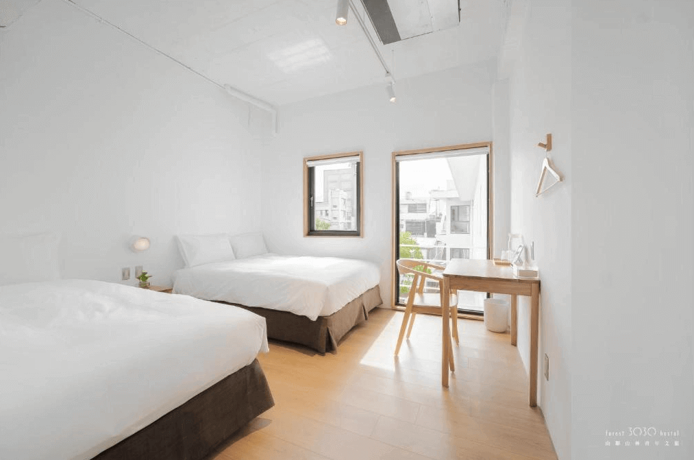 【玉里住宿】山鄰山林青年文旅的客房，擁有兩張白色床鋪，房間內光線充足，搭配木質地板和一張小書桌，窗外景色宜人，營造出舒適的住宿環境。