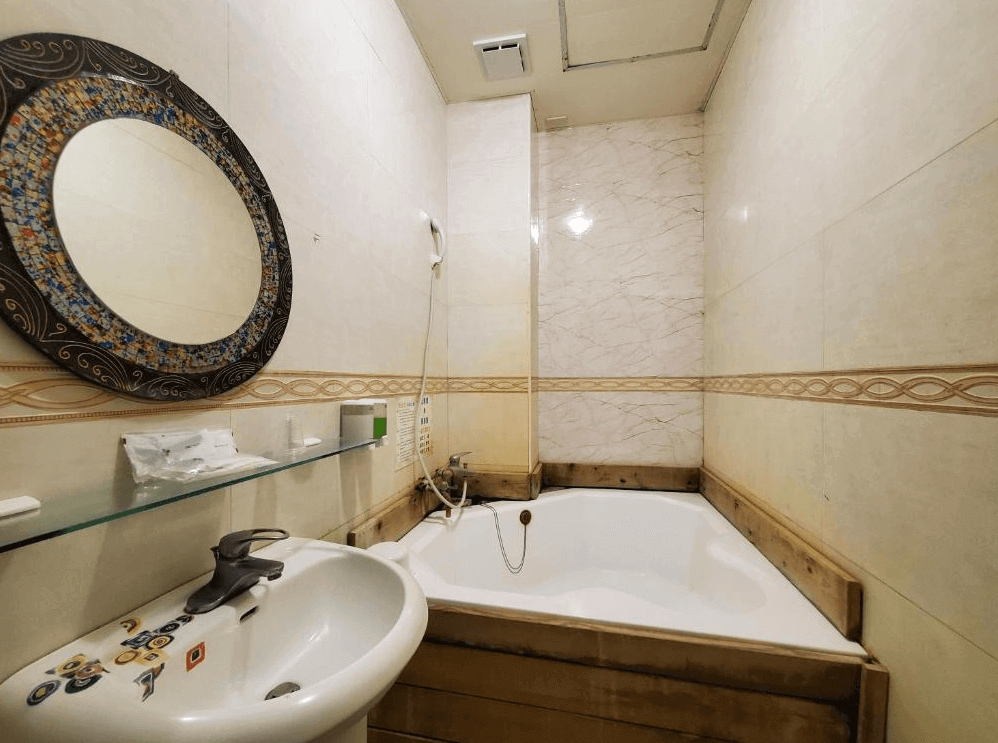 椰子林溫泉飯店的浴室，內有一個白色浴缸和洗手台。牆壁採用淺色瓷磚裝飾，圓形鏡子周圍有花紋裝飾，洗手台上方設有玻璃擱板，提供實用的收納空間。