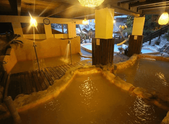 椰子林溫泉飯店的室內溫泉池，水池分成多個區域，提供不同的按摩體驗。周圍設有木製平台和噴泉，燈光柔和，營造出放鬆的氛圍，背景可見戶外的景色和休息區。