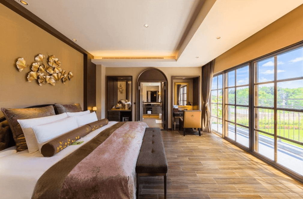 【花蓮寵物友善飯店】瑞穗天合國際觀光酒店的豪華雙人房，房間內有一張大床，床頭裝飾著金色的裝飾品。房間內設計典雅，配有大窗戶，提供充足的自然光。房間內還有一張長凳和一個小書桌，整體氛圍溫馨舒適。
