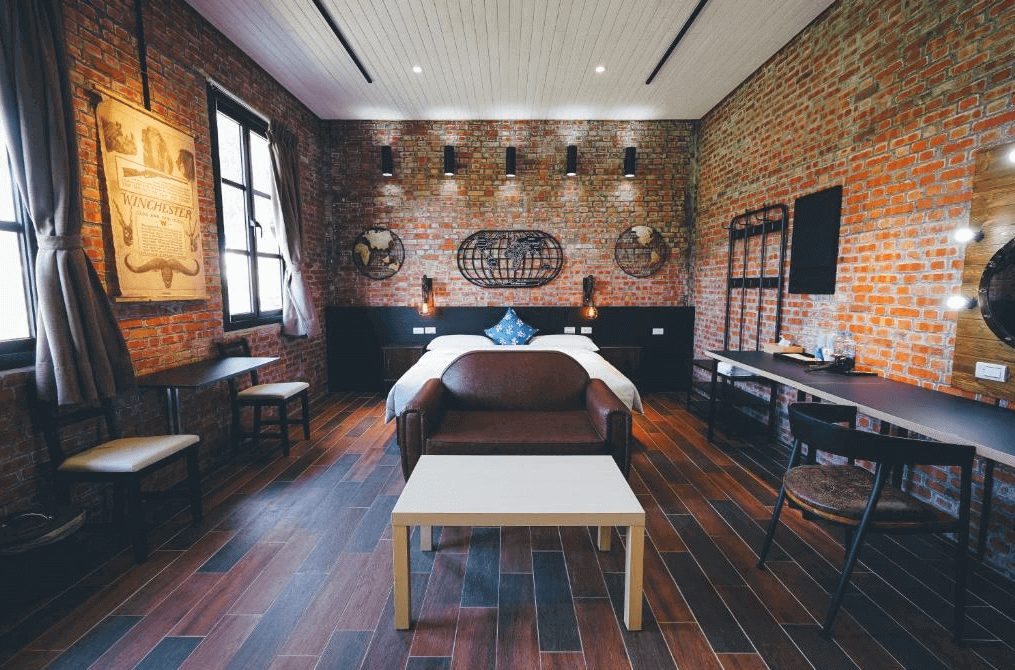 【美濃住宿】美濃煙城民宿的民宿房間，牆面採用紅磚裸露設計，房內配有雙人床、沙發和木製桌椅。窗戶旁掛有復古海報，房間內的燈光柔和，營造出溫馨的氛圍。
