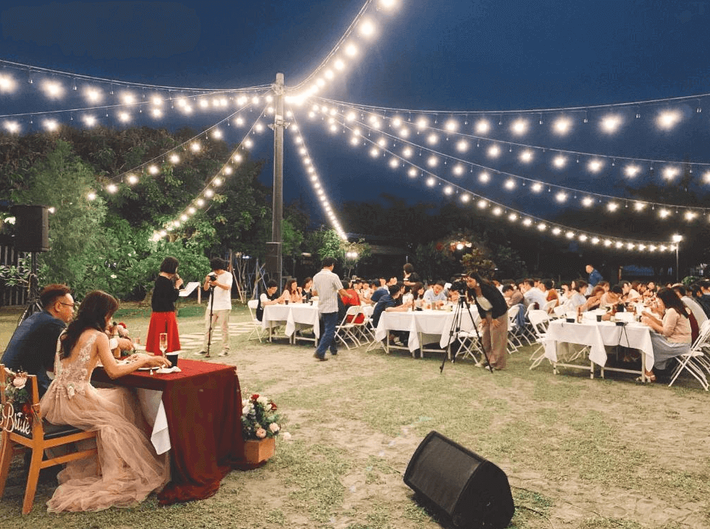 【美濃住宿】美濃煙城民宿的戶外活動場景，夜晚時分燈光環繞，氣氛浪漫。許多人在燈光下的庭院裡參加晚宴，背景是樹木和綠色植物，場地布置精緻，適合舉辦婚禮或大型聚會。