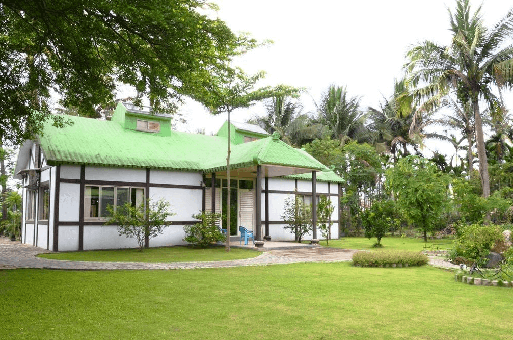 【美濃住宿】美濃百果園民宿，屋頂為綠色，周圍被綠色植物和樹木環繞。庭院內草坪整潔，空間寬敞，適合戶外活動和放鬆，是享受自然的理想場所。