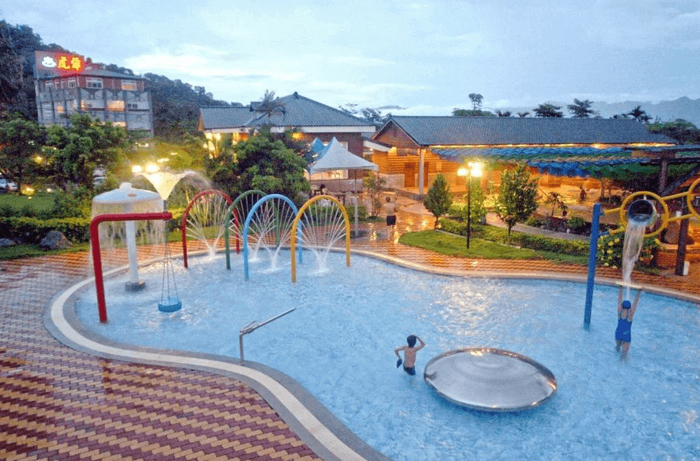 虎爺溫泉渡假莊園的兒童戲水池區域，池內設有各種彩色噴水設施和玩具。背景是度假村的建築和綠色植物，池邊鋪有防滑地磚，為小朋友提供安全的戲水環境，充滿歡樂氣氛。