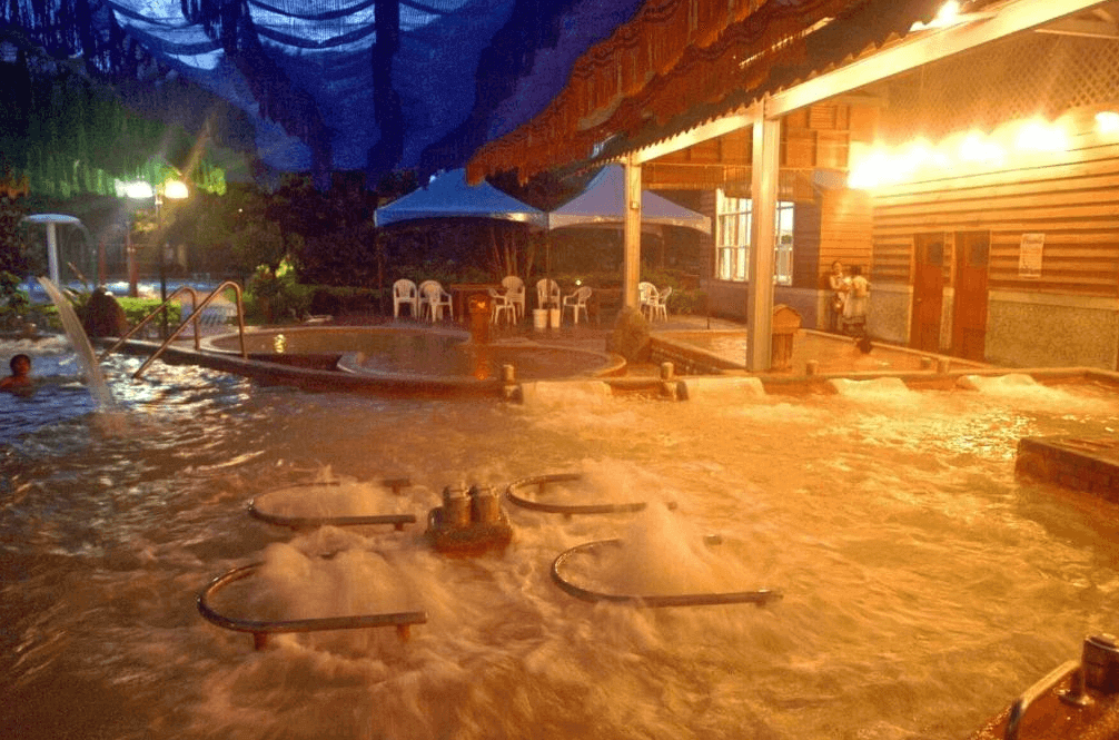 虎爺溫泉渡假莊園的溫泉水療區，夜晚拍攝，水池內有多個按摩噴頭，提供放鬆的水療體驗。周圍燈光柔和，營造出溫馨的氛圍，背景可見度假村的建築和休息區，讓住客在享受溫泉的同時放鬆心情。