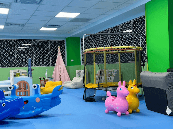 蜜滋賀溫泉飯店的室內兒童遊戲區，地面鋪有藍色軟墊，配有多種玩具和遊樂設施，如搖搖馬、滑梯和蹦床。牆壁為綠色，房間光線明亮，設有一個粉紅色的兒童帳篷，提供孩子們一個安全且有趣的遊戲環境。