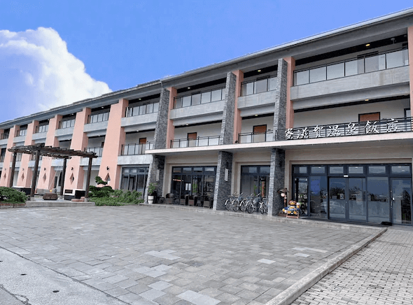 蜜滋賀溫泉飯店的外觀，建築設計現代，共有三層樓，每個房間都有獨立陽台。飯店入口寬敞，前方是鋪設整齊的石板路，兩旁有綠色植栽，營造出舒適的迎賓氛圍。