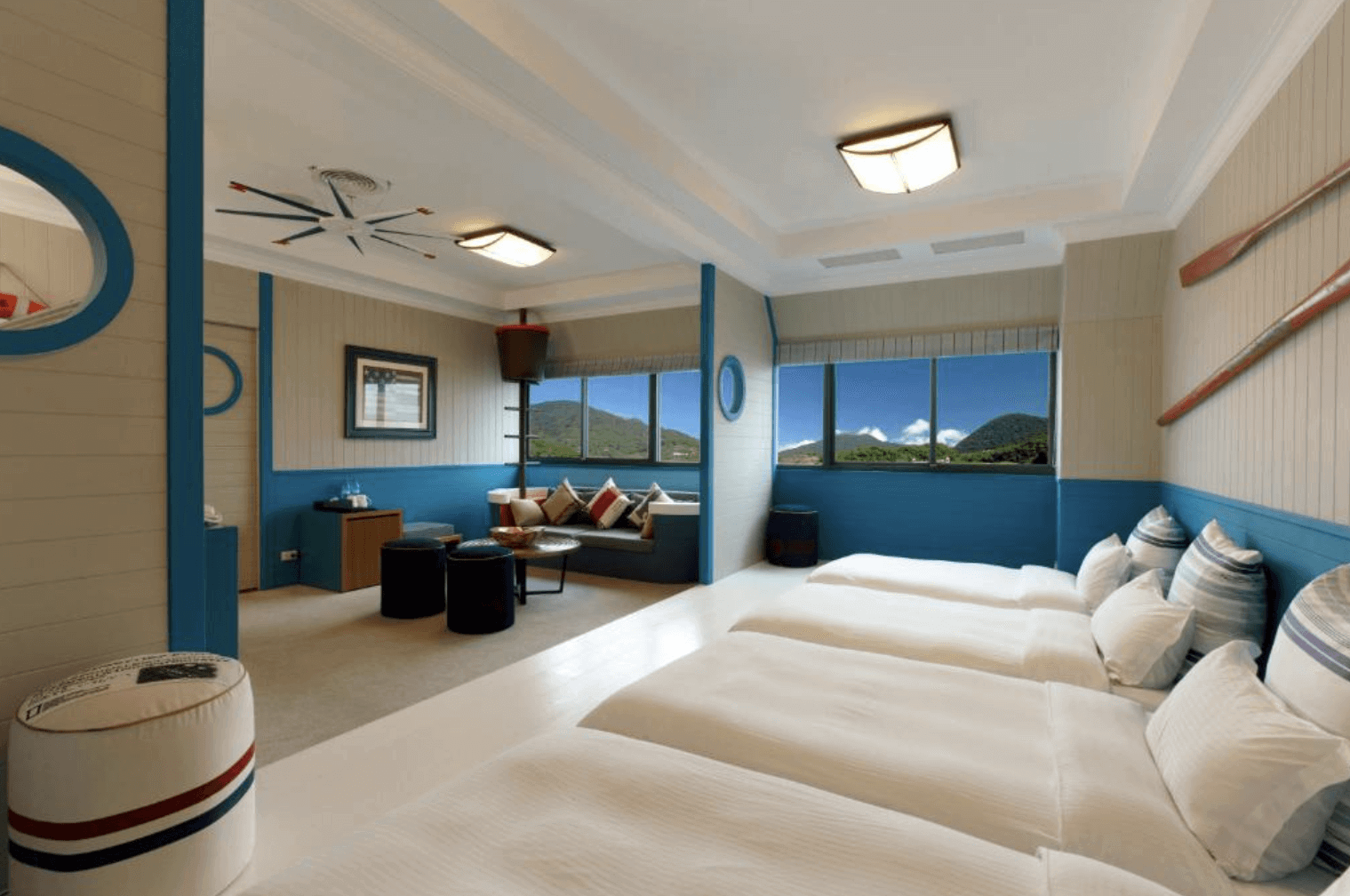 "台北親子飯店"【陽明山親子住宿】陽明山天籟渡假酒店的客房，房間以藍白色調為主，給人清新舒適的感覺。客房內設有三張床，整齊排列，提供足夠的睡眠空間。房間一側有大窗戶，可以看到窗外的美麗山景，讓人感受到大自然的靜謐與美好。房間還設有一個小型休息區，配有舒適的沙發和茶几，非常適合放鬆和享受愜意的時光。天花板上裝飾有創意十足的吊燈，牆上也有一些裝飾物，如槳和圓形鏡子，增添了房間的獨特性和趣味性。