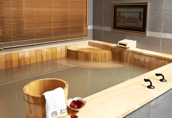 陽明山家新休閒會館木質溫泉浴池：一間設有大型木質溫泉浴池的浴室，旁邊擺放著木質水桶和毛巾，窗戶上有竹製百葉窗。浴池內水質清澈，整體設計典雅且放鬆