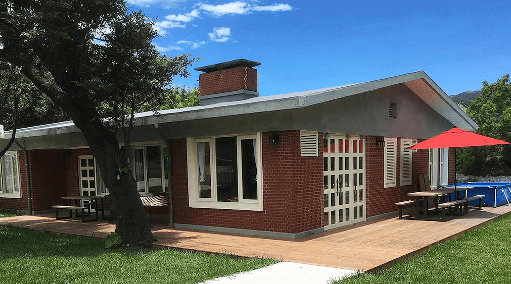 陽明山美國渡假村外觀：一棟紅磚建築的平房，屋頂上有煙囪，房子周圍有綠色草坪和樹木。戶外有木質平台，設有木製桌椅和紅色遮陽傘，背景是晴朗的藍天白雲