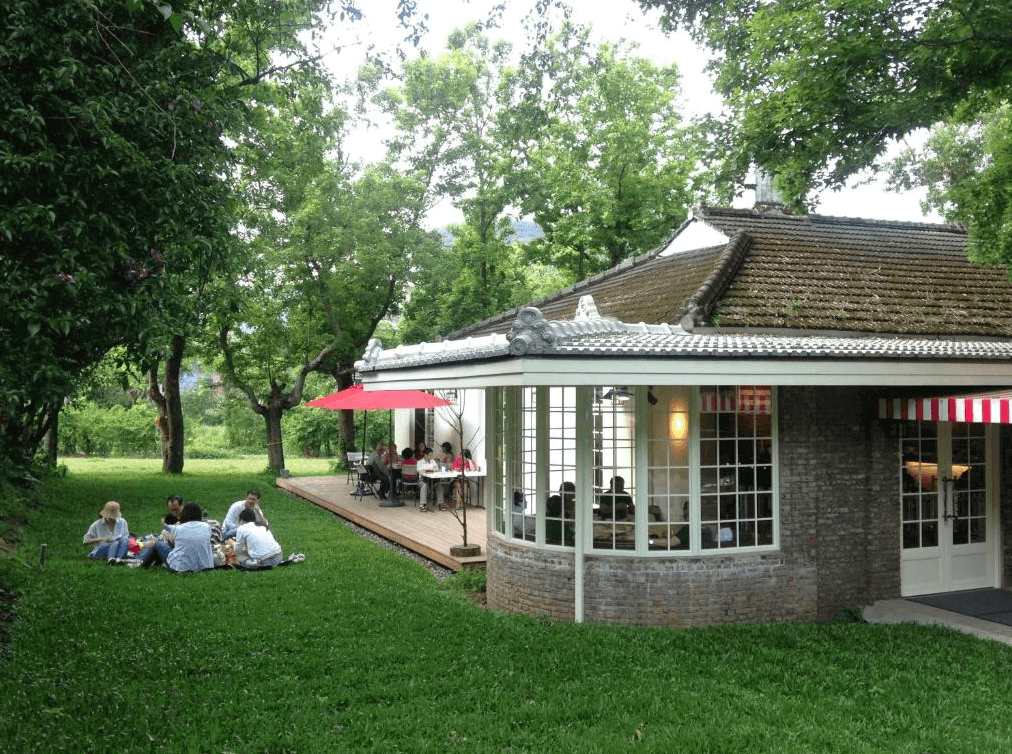 陽明山美國渡假村戶外活動區：一群人在綠色草坪上進行野餐和戶外活動。背景是一棟帶有大窗戶和白色門的建築，屋頂上有傳統的瓦片，戶外有木製平台和紅色遮陽傘