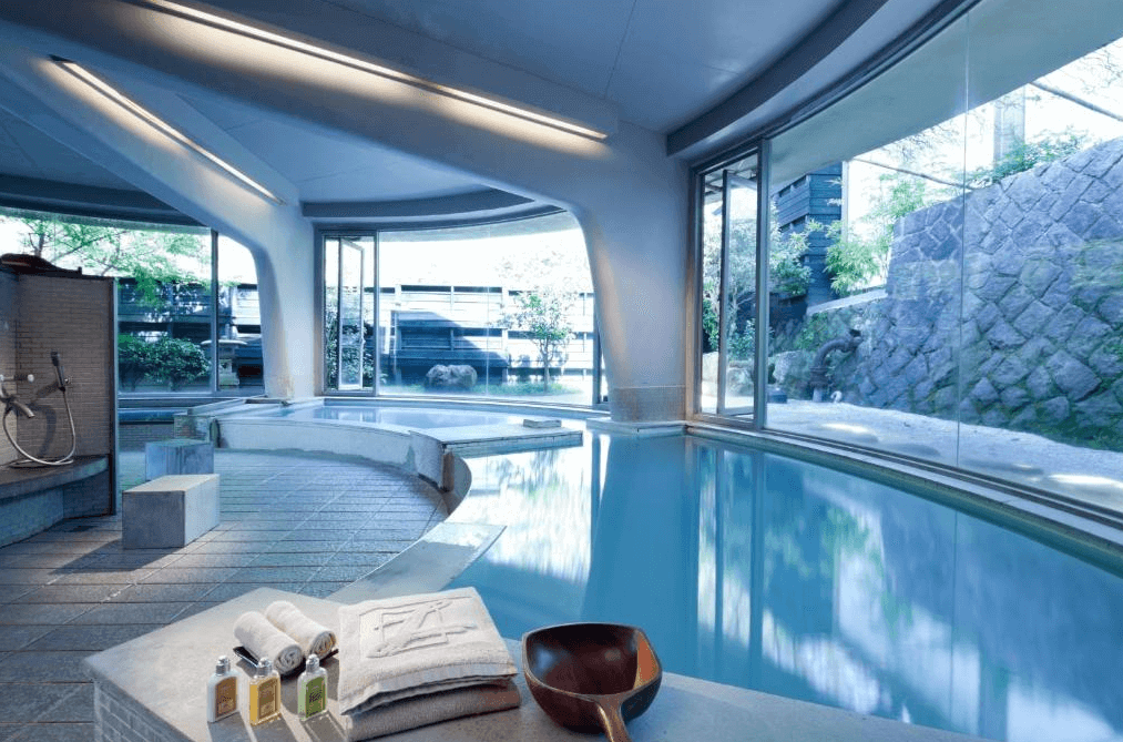 雀客藏居臺北陽明山室內溫泉：一個設有大型落地窗的室內溫泉池，窗外可見綠意盎然的景色。溫泉區域設計現代，提供毛巾、洗漱用品和木質水勺，營造出舒適放鬆的氛圍
