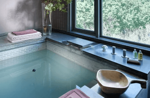 雀客藏居臺北陽明山私人溫泉：一間設有大窗戶的私人溫泉浴室，窗外景色優美。浴室內設有石製浴缸，旁邊擺放著毛巾和洗漱用品，整體設計簡約且高雅