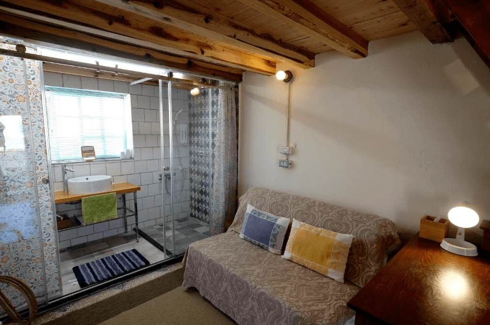 【美濃住宿】香蕉與黑膠三合院民宿房間，房內有一個木製的閣樓，樓梯通往上層。下層的沙發區域配有柔和的燈光，並有一間配有淋浴間和洗手台的浴室，浴室內裝有白色瓷磚牆面，並用花紋窗簾隔開。